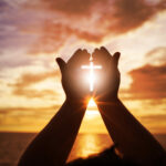 Two human hands forming a Christian cross with the sun shining through.