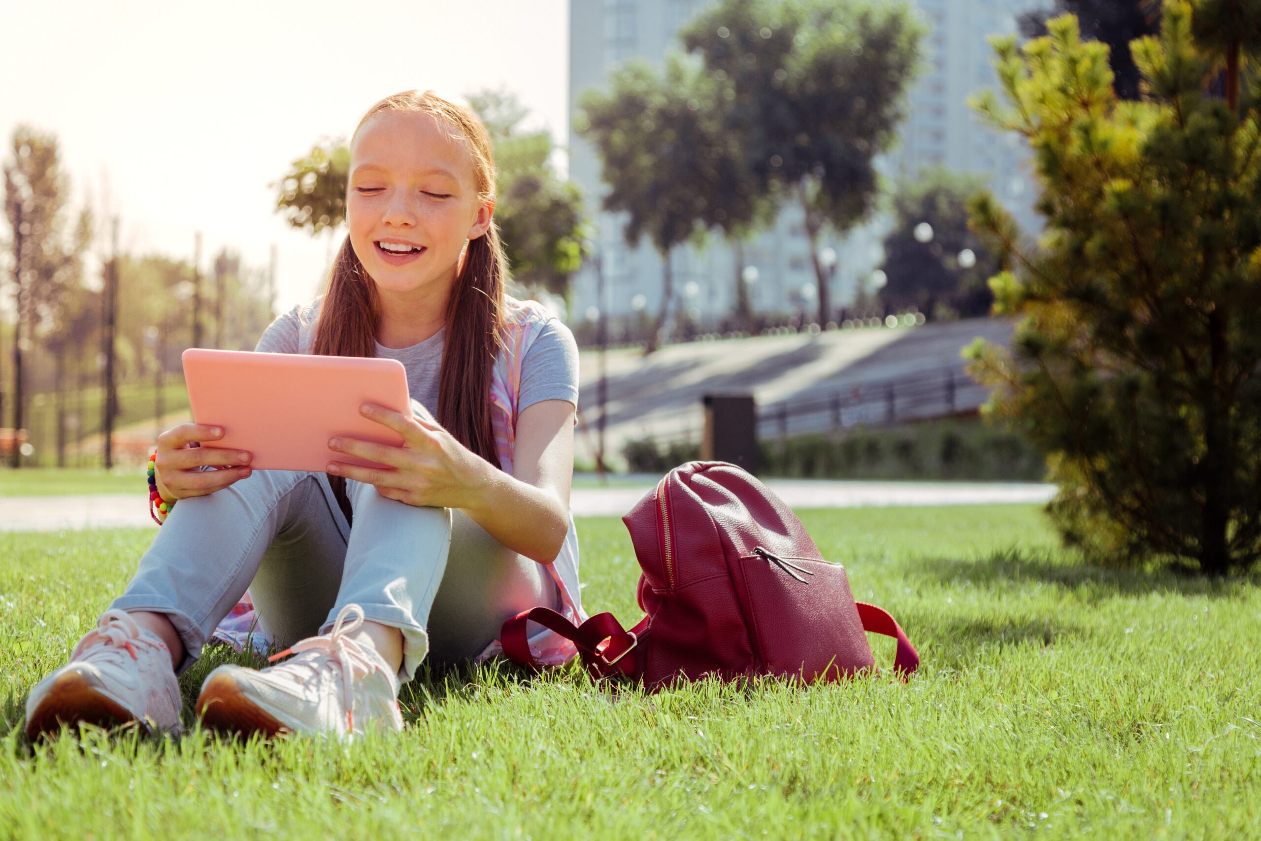Green grass. Cheerful girl sitting on grass while staring at her tablet