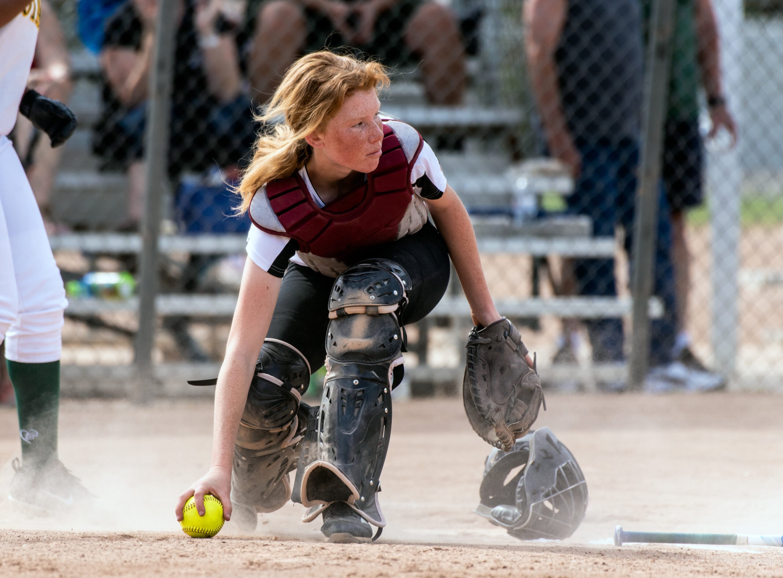 Skilled softbaall catcher with red hair and protective gear gaining a grip on the loose ball while looking up field.