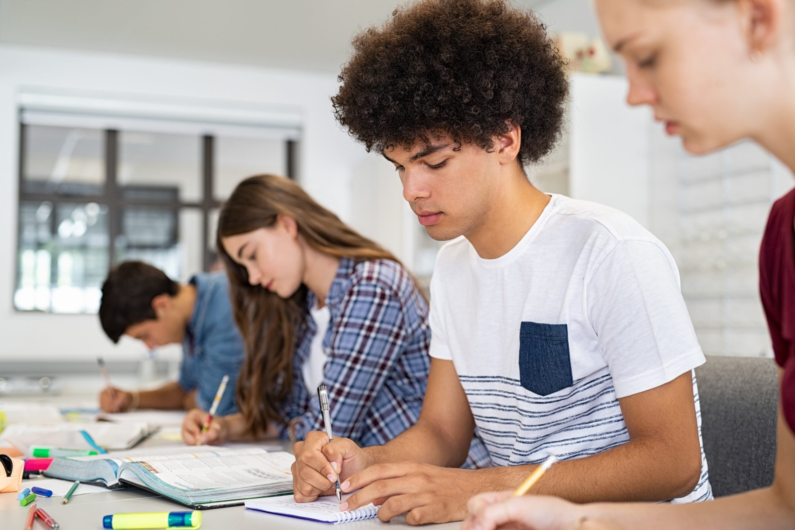 Concentrated young man studying in classroom with friends sitting in a row. College student concentrating on studies while taking notes in university library. African american high school guy preparing exam with classmates in class.