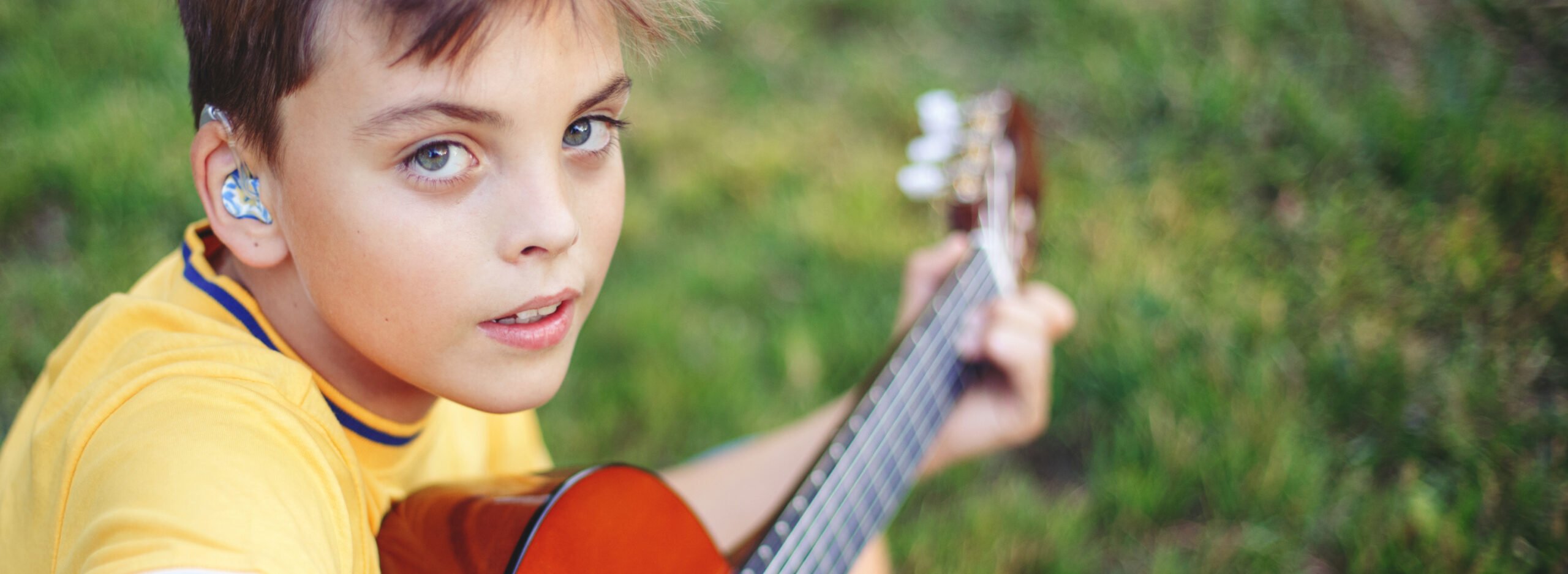 Hard of hearing preteen boy playing guitar outdoor. Child with hearing aids in ears playing music and singing song in park. Hobby art activity for children kids. Web banner header.