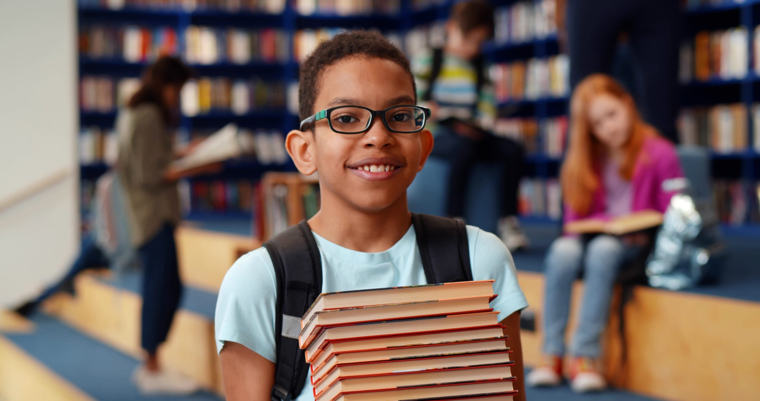 Middle eastern boy holding stack of books against multi colored bookshelf in library. Portrait of happy african schoolboy with textbooks smiling at camera in school library