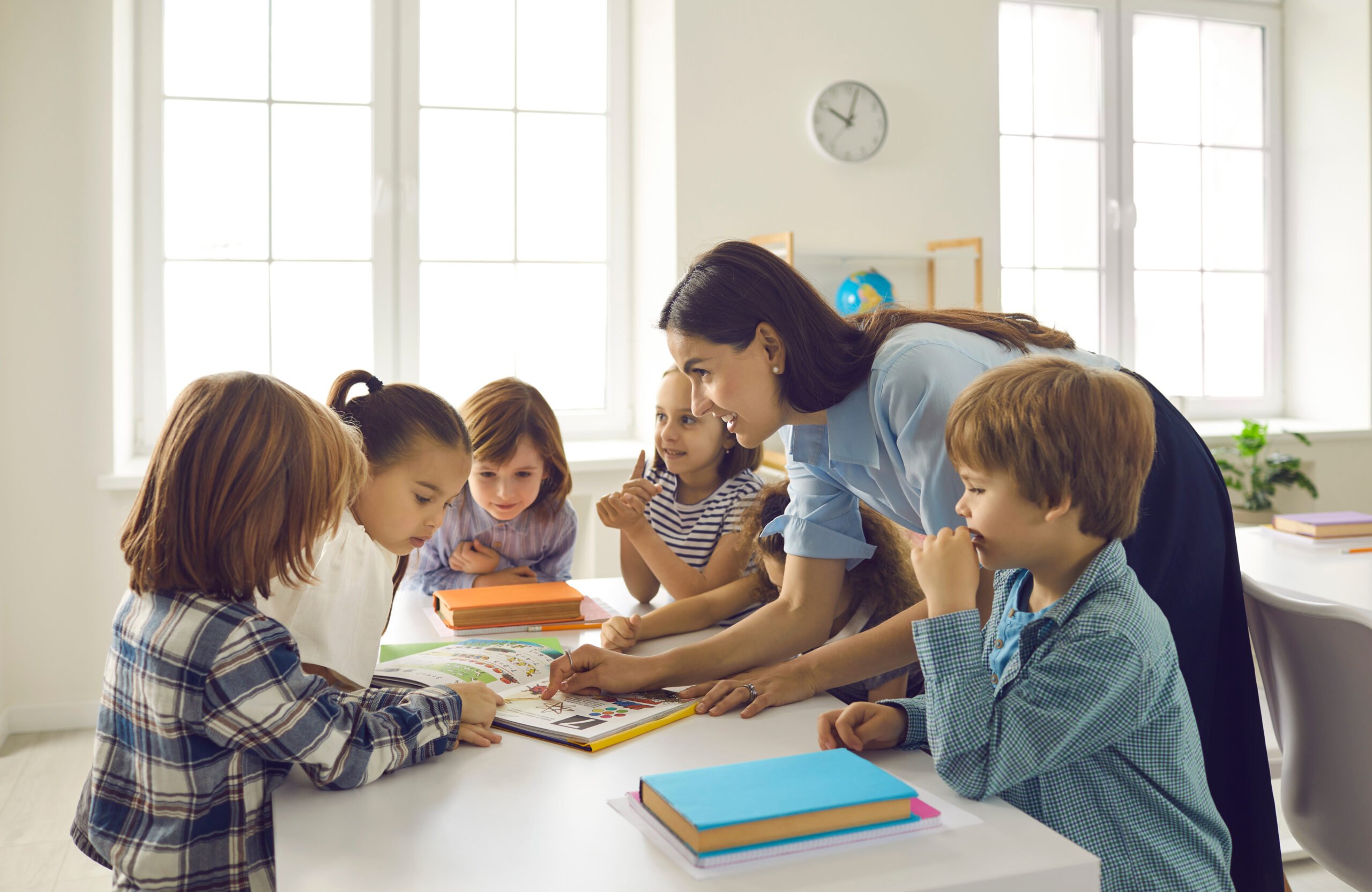 Happy teacher and children reading book together. Group of school kids gather around table in classroom to listen to interesting story told by teacher. Elementary students learning new things in class