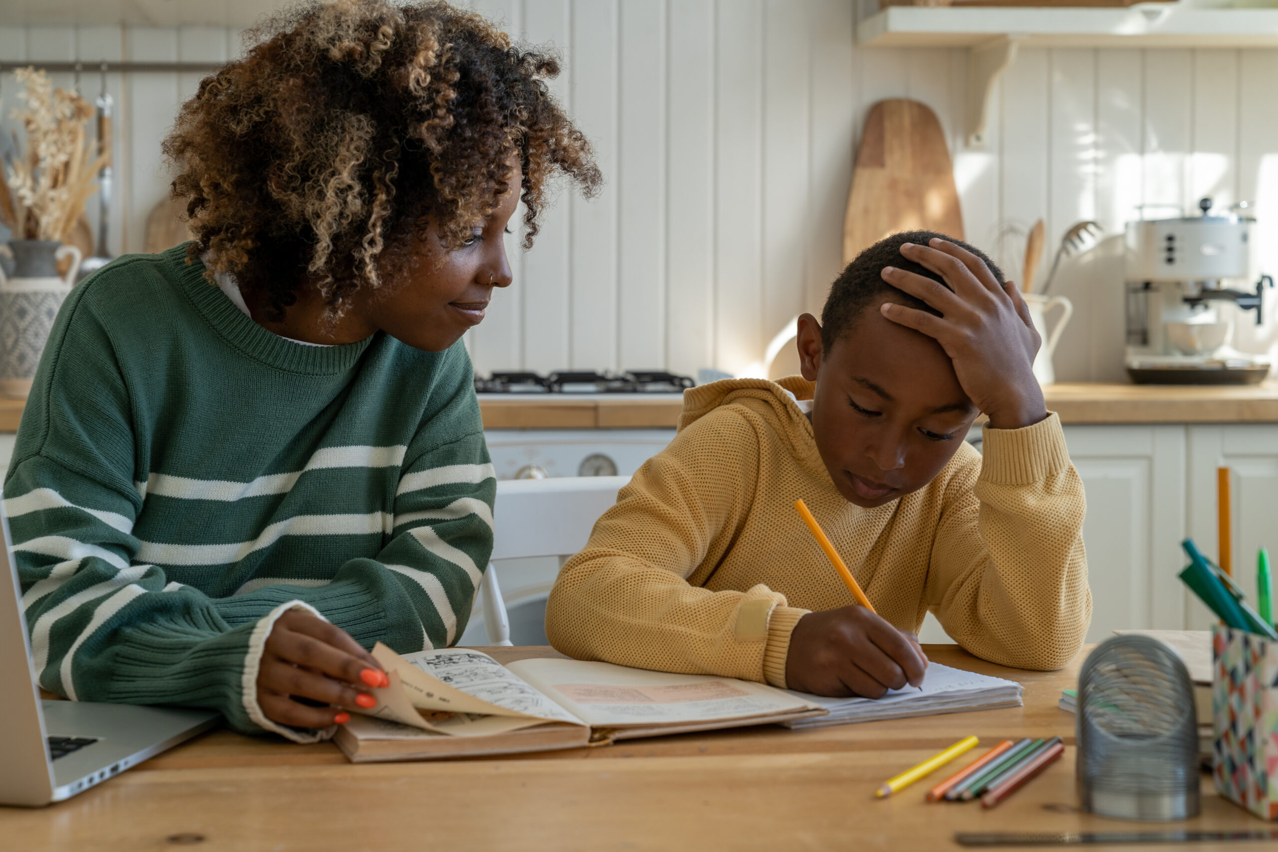 Young African American woman homeschool mom helping son with homework, mother and child studying at home together. Parent sitting at table with kid checking assignment. Parenthood and homeschooling