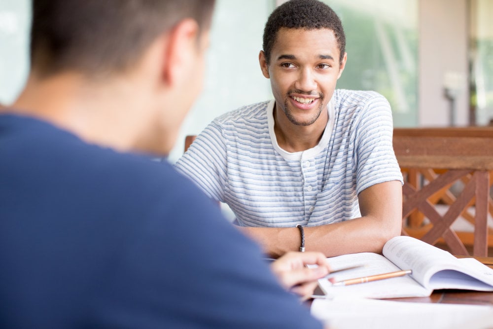 Cheerful male student discussing project with groupmate and looking at him. Positive young man sitting at table and talking about homework. Private tutor concept