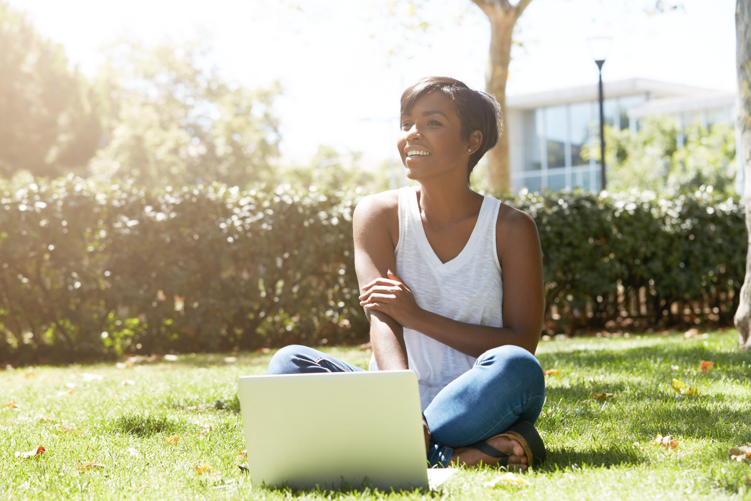 Happy and cheerful black female with short haircut sitting on the grass, smiling to her friends approaching her. Young African student using her notebook outdoor, resting after a hard day at college