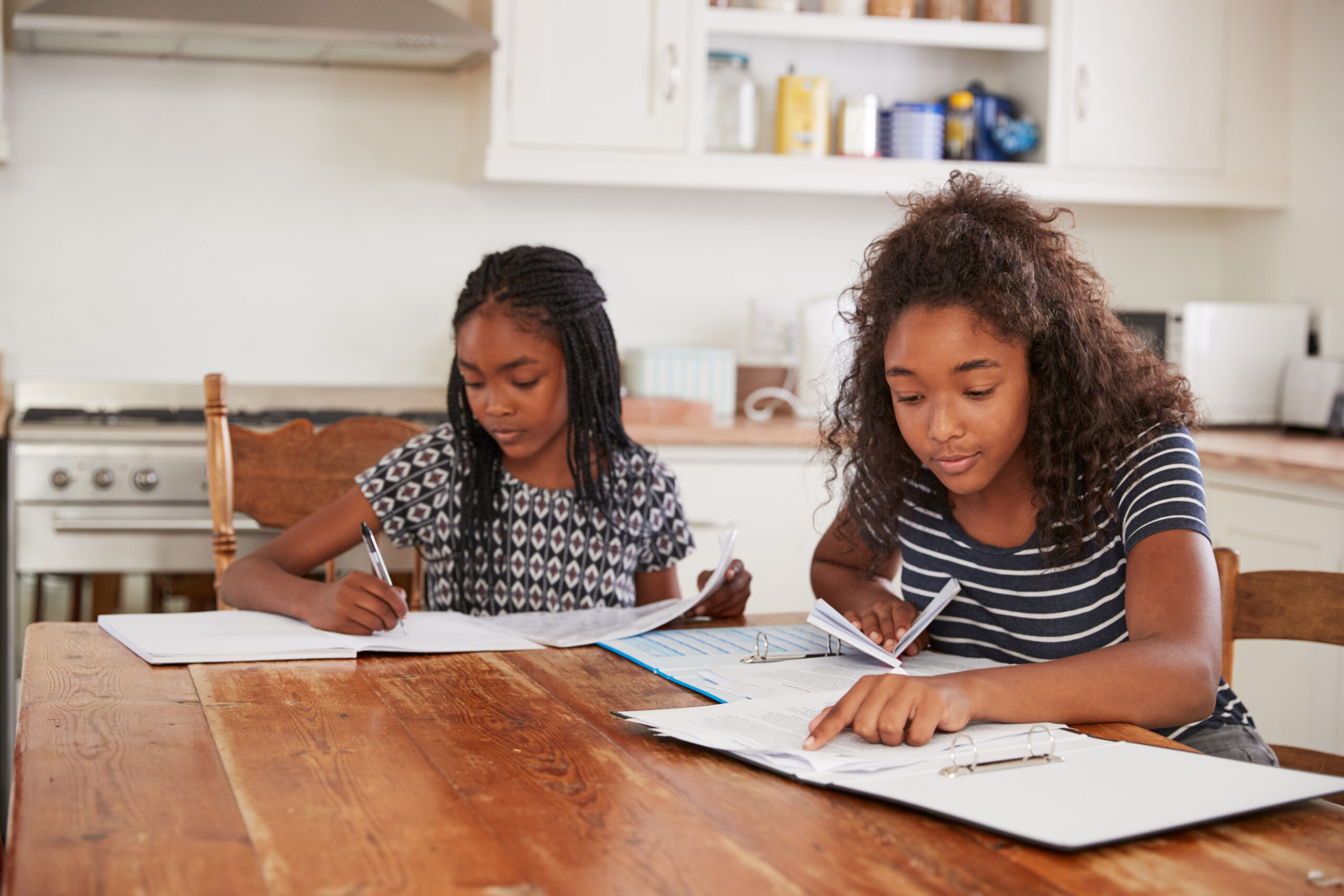 Two Sisters Sitting At Table In Kitchen Doing Homework