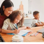 A parent sits with her children at a table where they are learning for school.